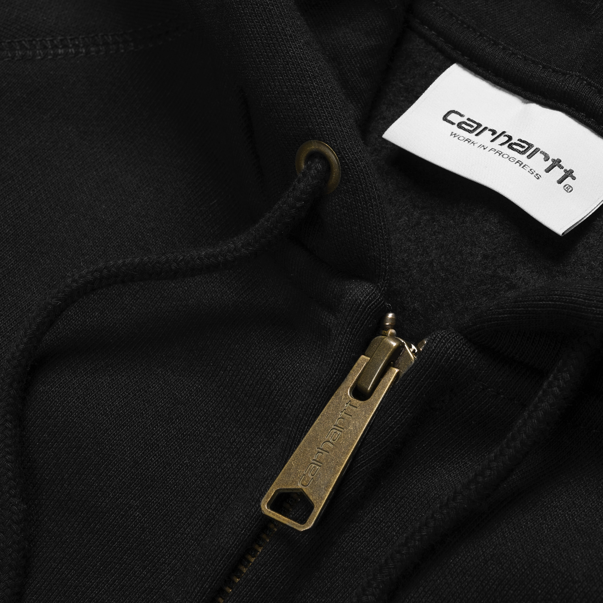 Carhartt WIP Hooded Chase Jacket em preto com logo bordado na manga em dourado. Foto de detalhe do fecho.